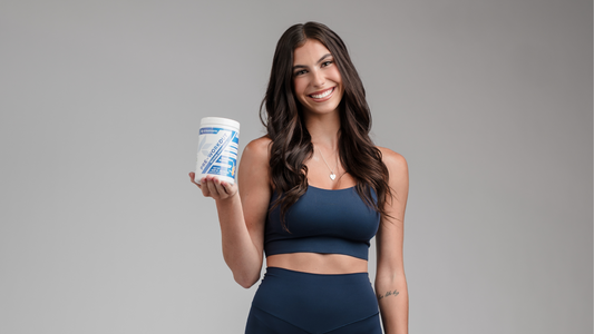 Athletic woman in navy workout gear holding a container of K3 Nutriments Pre-Workout Tropical Sunrise, smiling confidently in a studio setting.