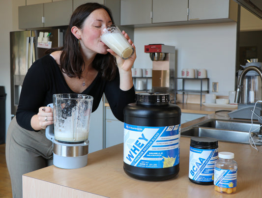Holly, a young woman and K3 Nutriments customer, preparing a vanilla whey protein shake with K2 + D3 supplements visible on the counter—representing a strong supplement stack for young women.